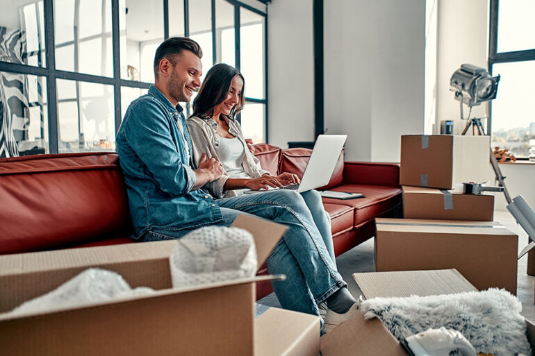 Young couple sitting on floor together using computer wireless internet while moving into new home. Moving, buying a house, apartment concept.